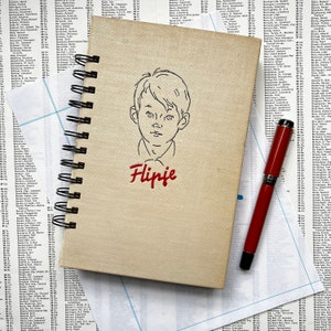 Pode incluir: Um caderno bege com uma encadernação em espiral preta. O caderno tem um desenho de linha de um rosto de menino e a palavra "Flipje" escrita em itálico vermelho.