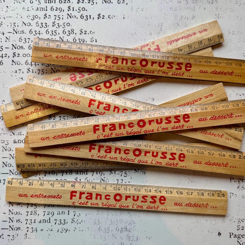 Puede incluir: Una pila de reglas de madera con letras rojas y doradas. Las reglas est&aacute;n etiquetadas "Francorusse" y dicen "c'est un r&eacute;gal que l'on sert... au dessert..." Las reglas miden 30,5 cm de largo.
