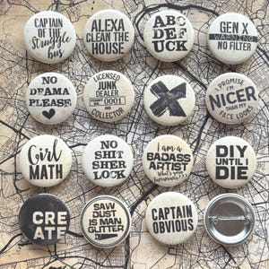 Op de afbeelding: Een verzameling ronde, gebroken witte buttons met zwarte tekst. De buttons tonen verschillende zinnen zoals "Girl Math", "No Drama Please" en "Captain Obvious". Sommige buttons hebben grafische elementen zoals een hart of een "X".