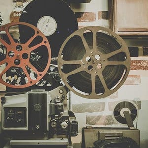 May include: Vintage film projector with two large film reels, one red and one black, and a smaller black reel. The projector is on a wooden surface with a brick wall in the background.