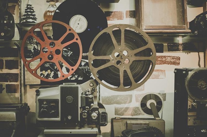 May include: Vintage film projector with two large film reels, one red and one black, and a smaller black reel. The projector is on a wooden surface with a brick wall in the background.