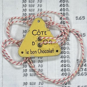 Könnte beinhalten: Ein gelbes dreieckiges Etikett mit dem Text "CÔTE D'OR le bon Chocolat" in schwarzer Schrift. Das Etikett ist an einer rot-weiß gestreiften Schnur befestigt. Der Hintergrund zeigt ein Kassenbuch mit Daten und Zahlen.