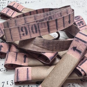 Könnte beinhalten: Eine Ansammlung von aufgerollten rosa Stoffbändern mit schwarzen Aufdrucken, die an ein Maßband erinnern. Jedes Band ist mit einem braunen Papierstreifen fixiert und zeigt Zahlen sowie vertikale Linien mit nach oben gerichteten Pfeilen.