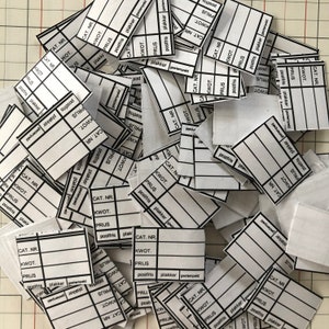 Op de afbeelding: Een stapel witte papieren etiketten met zwarte tekst. De etiketten zijn voor een productcatalogus en bevatten velden voor het artikelnummer, de hoeveelheid, de prijs, de verzendkosten en of het artikel verpakt of gestempeld is.