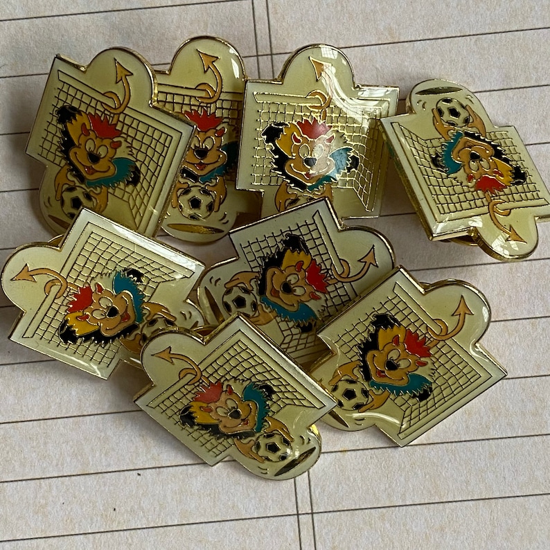 May include: A set of nine gold-colored metal pins featuring a cartoon character with a soccer ball and a goal net. The pins are in various poses and have a white background.