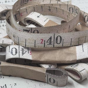 Può includere: Una collezione di nastri di misurazione arrotolati in carta color bianco sporco con segni e numeri stampati in nero. Ogni nastro è fissato con una fascetta di carta marrone. I numeri visibili includono 40 e 2, con frecce nere direzionali.
