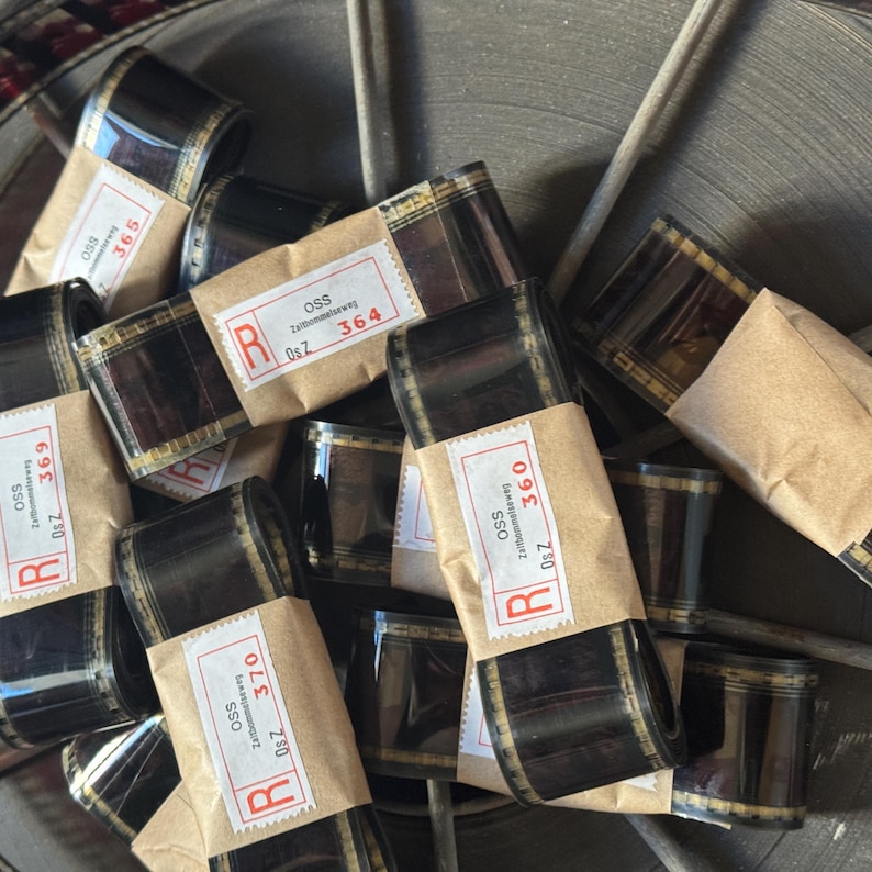 May include: A close-up of several rolls of 35mm film, each wrapped in brown paper with a red label. The labels have the text "OSS Zaltbommelseweg Os Z" and a number, such as "364" or "369".