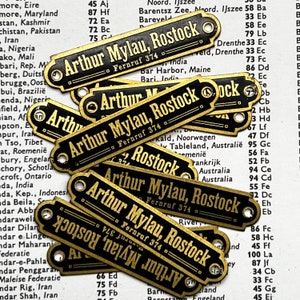May include: A close-up of several brass tags with black lettering. The tags are rectangular and have rounded corners. The text on the tags reads "Arthur Mylau, Rostock" and "Fernruf 374".