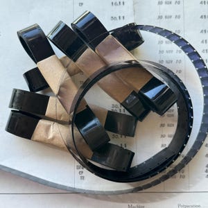 May include: A collection of vintage black film strips, some coiled and others wrapped in brown paper. The film strips are arranged on a paper surface with printed text and numbers, including "30 NOV 70" and "16.11".