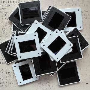 May include: A collection of vintage 35mm slide mounts, both black and white, are scattered across a textured surface. The slides have a rectangular viewing area and are made of plastic. The image suggests a theme of photography and retro media.