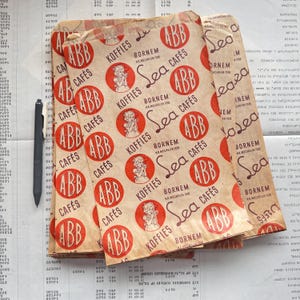 May include: Vintage paper coffee bags with a repeating pattern of red circles with the letters "ABB" and the words "Sea Caf&eacute;s" and "Koffies". The bags are stacked on a ledger sheet with a pen.
