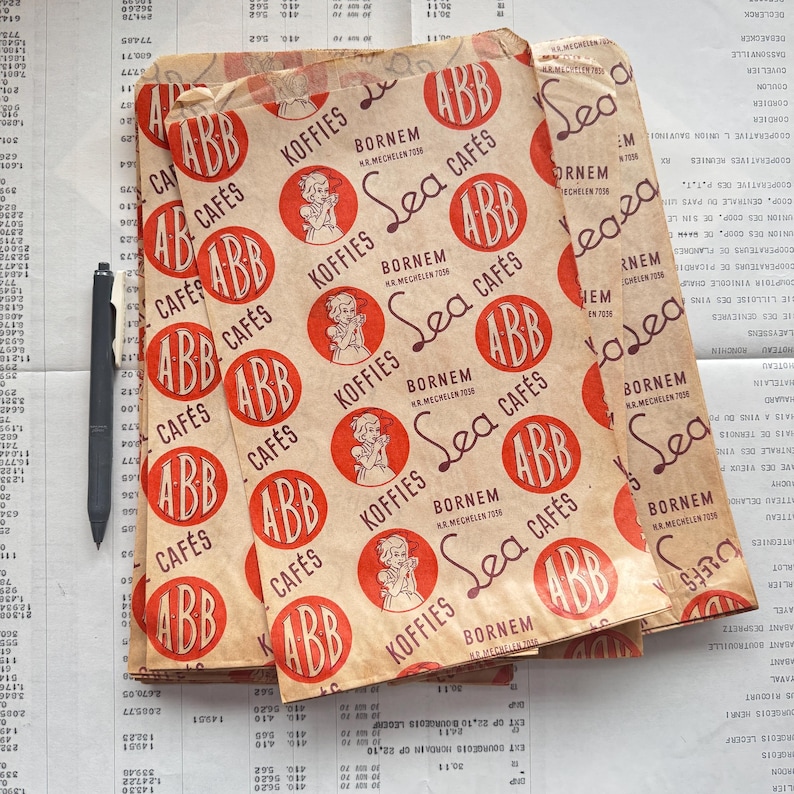 May include: Vintage paper coffee bags with a repeating pattern of red circles with the letters "ABB" and the words "Sea Caf&eacute;s" and "Koffies". The bags are stacked on a ledger sheet with a pen.