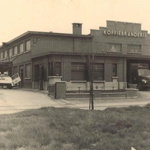 May include: Vintage black and white photograph of a brick building with the text "KOFFIEBRANDERIJ" above the entrance. Several vehicles, including a truck and a van, are parked in front of the building. The image has a classic, historical feel.