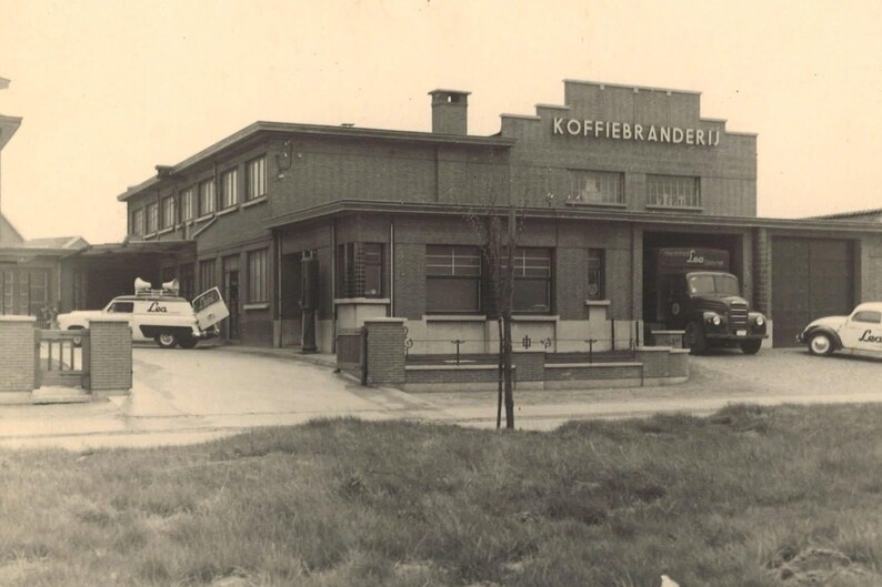 May include: Vintage black and white photograph of a brick building with the text "KOFFIEBRANDERIJ" above the entrance. Several vehicles, including a truck and a van, are parked in front of the building. The image has a classic, historical feel.