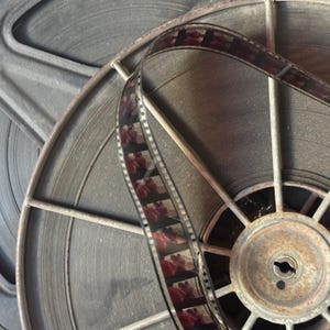 May include: Close-up of a vintage film reel with a strip of film running through it. The reel is made of metal and has a rusty appearance. The film is black and white and has a grainy texture.