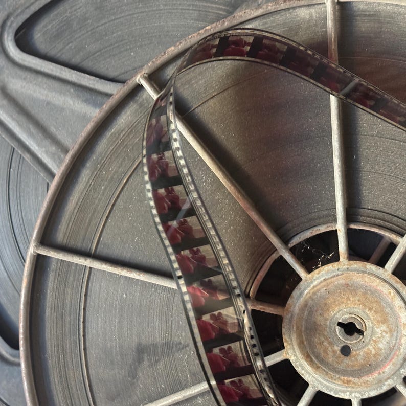 May include: Close-up of a vintage film reel with a strip of film running through it. The reel is made of metal and has a rusty appearance. The film is black and white and has a grainy texture.