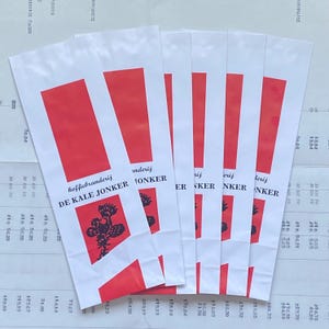 May include: Several white paper bags with a red rectangular design at the top and a vertical red stripe. The bags have text and a floral graphic in black. The text reads "koffiebranderij DE KALE JONKER".