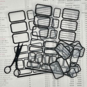 May include: A sheet of clear, transparent labels with black outlines. The labels are rectangular with rounded corners and have lines inside. The labels are arranged in rows and columns. A pair of black scissors is lying on top of the labels.
