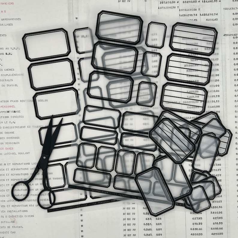 May include: A sheet of clear, transparent labels with black outlines. The labels are rectangular with rounded corners and have lines inside. The labels are arranged in rows and columns. A pair of black scissors is lying on top of the labels.