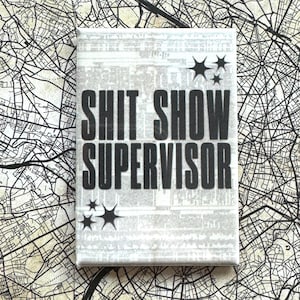 Può includere: Stampa su tela rettangolare con la scritta "SHIT SHOW SUPERVISOR" in grassetto nero. Lo sfondo è una stampa di giornale grigia sbiadita. Grafiche a forma di stella nera negli angoli. La tela è posta su uno sfondo di mappa.