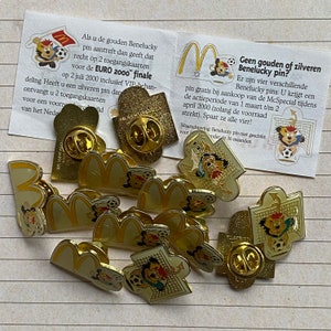 May include: A collection of 11 gold and yellow metal pins featuring the McDonald's golden arches and a cartoon character. The pins are on a white and gray striped surface. The text on the card reads: "Geen gouden of zilveren Benelucky pin?"