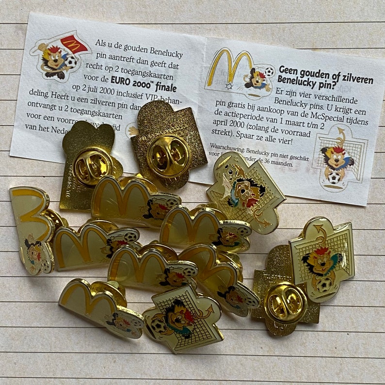 May include: A collection of 11 gold and yellow metal pins featuring the McDonald's golden arches and a cartoon character. The pins are on a white and gray striped surface. The text on the card reads: "Geen gouden of zilveren Benelucky pin?"