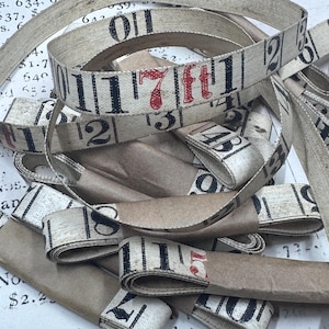 Può includere: Una collezione di nastri in tessuto color bianco sporco stampati con segni numerici neri e testo rosso che recita 7ft. I nastri sono arrotolati e fissati con fascette di carta marrone, caratterizzati da un design vintage con numeri.