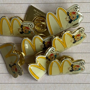 May include: A collection of seven yellow and white enamel pins featuring the McDonald's golden arches logo and cartoon characters playing soccer. The pins are in various sizes and feature different characters.