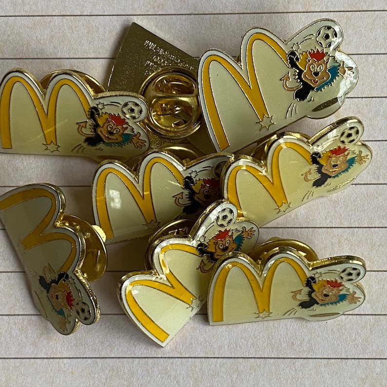 May include: A collection of seven yellow and white enamel pins featuring the McDonald's golden arches logo and cartoon characters playing soccer. The pins are in various sizes and feature different characters.