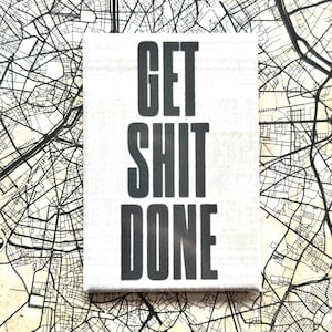 Op de afbeelding: Een witte canvas met de tekst "GET SHIT DONE" in grote, zwarte hoofdletters. De achtergrond is een zwart-witte kaart.