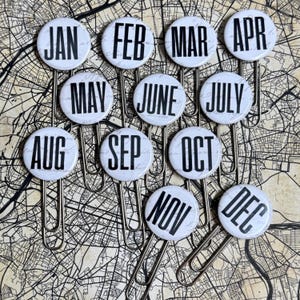 May include: Twelve white paper clips, each with a circular design displaying a month of the year in bold black text. The clips are arranged on a map-like background, creating a calendar-themed aesthetic.