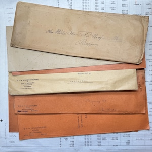 Op de afbeelding: Een verzameling vintage enveloppen in verschillende maten en kleuren, waaronder bruin en oranje. Handgeschreven adressen en tekst zijn zichtbaar op de enveloppen, samen met wat gedrukte tekst. De enveloppen zijn op elkaar gestapeld.