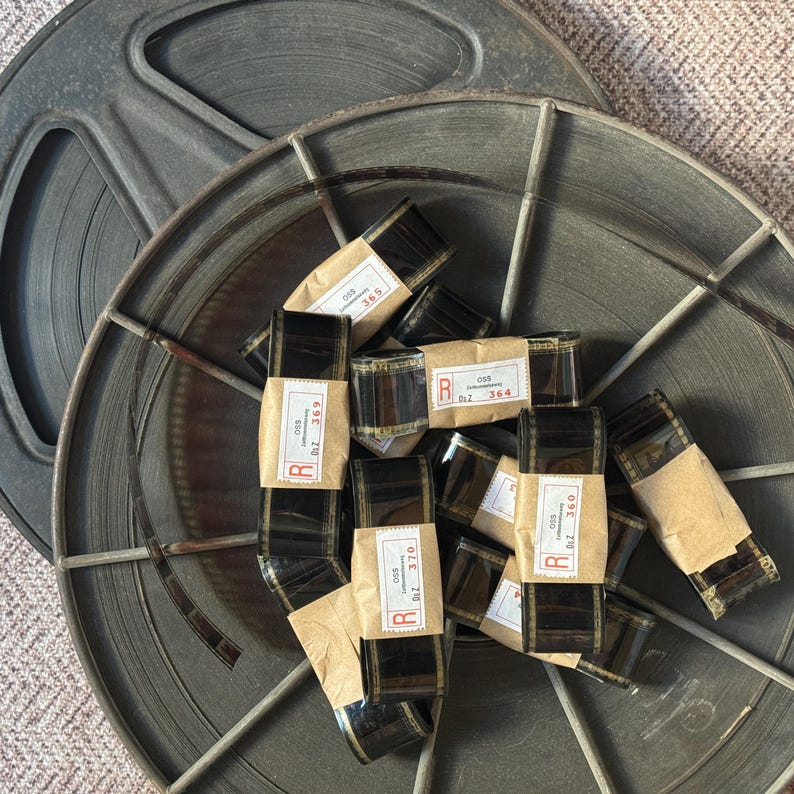 May include: A close-up of several rolls of film labeled with the letters "R" and "OSS" and numbers. The film is sitting inside a metal film reel.