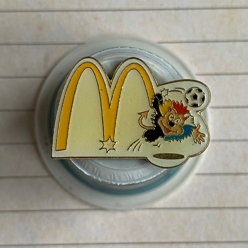 May include: A gold-colored pin featuring the McDonald's golden arches with a cartoon character kicking a soccer ball. The character is wearing a blue and black jersey with a red and yellow mohawk.