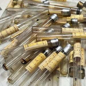 Könnte beinhalten: Nahaufnahme einer Sammlung von Vintage-Parfümfläschchen. Die durchsichtigen Glasröhrchen haben silberne Kappen und gealterte Papieretiketten mit verblasstem Text. Die Fläschchen sind in einem Haufen angeordnet.