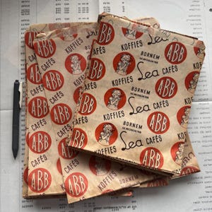 May include: Several vintage paper coffee bags are stacked, featuring a repeating pattern of red circles with a woman illustration and the text "ABB Caf&eacute;s" and "Koffies". The bags have a distressed, aged appearance.