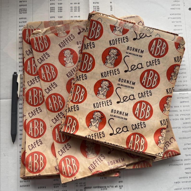 May include: Several vintage paper coffee bags are stacked, featuring a repeating pattern of red circles with a woman illustration and the text "ABB Caf&eacute;s" and "Koffies". The bags have a distressed, aged appearance.