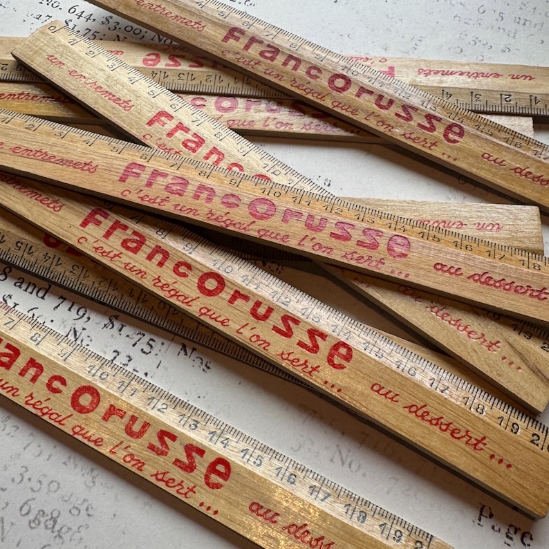 Puede incluir: Una pila de reglas de madera con el texto "Francorusse c'est un r&eacute;gal que l'on sert..." impreso en rojo en cada regla. Las reglas est&aacute;n marcadas en cent&iacute;metros y pulgadas.