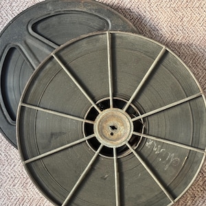 May include: Two vintage film reels with metal spools and black film. The reels are stacked on top of each other.