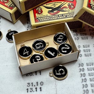 Op de afbeelding: Een verzameling zwart en goudkleurige bedels met cijfers en de letter "A" in een kleine kartonnen doos. De bedels hebben een gouden rand en een kleine lus om te bevestigen. Lucifersdoosjes zijn op de achtergrond te zien.