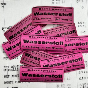 May include: A collection of pink paper labels with the text "Mohren-Apotheke Wasserstoff E. & L. Schurer -:- Bad Wimpfen Fernruf 747" printed on them.