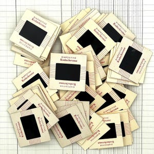 May include: A collection of vintage Kodachrome slides. Each slide is in a cream-colored cardboard frame with the text "DIAPOSITIVE Kodachrome" in red. The slides have a black rectangular viewing area and are arranged on a white grid.