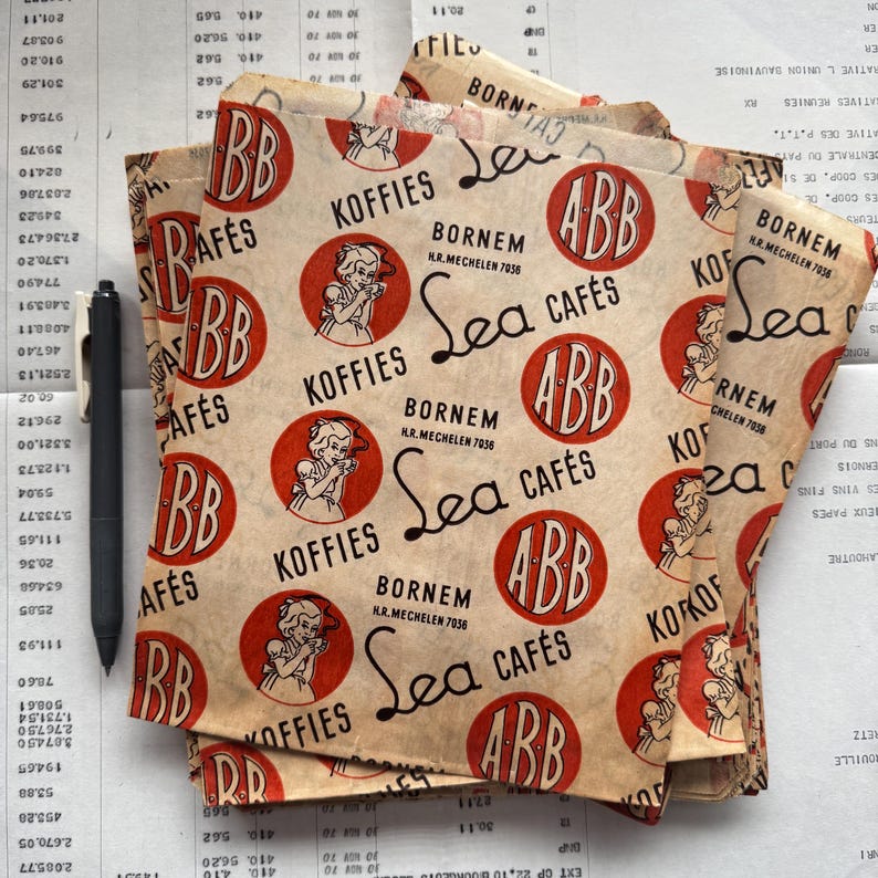 May include: A stack of vintage paper coffee bags with a repeating design of red circles with the letters "ABB" and "BB", the words "Koffies" and "Sea Caf&eacute;s", and an illustration of a woman. A black pen rests on the stack.