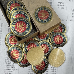 Op de afbeelding: Een verzameling ronde, goudkleurige etiketten met een blauw en rood ontwerp. De etiketten bevatten de tekst "Bowers Pottery Co. Mannington, W.VA." en een gestileerd "B"-logo. De etiketten zijn gerangschikt in een doos en op een tafel.