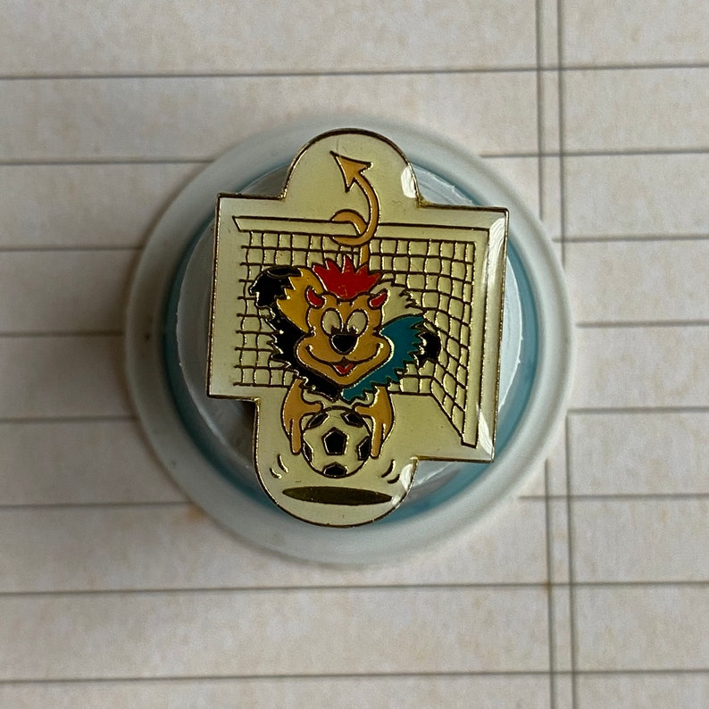 May include: A gold-colored pin with a cartoon devil character holding a soccer ball in front of a soccer goal. The devil has red hair, a yellow and black striped tail, and a blue and white striped shirt.