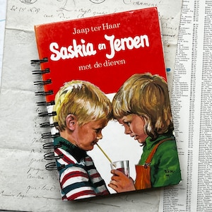 Può includere: Un quaderno a spirale rosso con un'illustrazione colorata di due bambini sulla copertina. Il titolo del libro è "Saskia en Jeroen met de dieren" di Jaap ter Haar.
