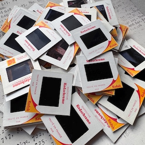 Può includere: Un mucchio di supporti per diapositive Kodak Kodachrome vintage. Ogni supporto è bianco con un'apertura rettangolare nera e un logo Kodak giallo e rosso. Il testo "TRAITE EN FRANCE PAR" è stampato sui supporti.