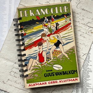 Pode incluir: Caderno com espiral com uma ilustração vintage de um clube de remo. A capa apresenta o texto "DE KANOCLUB" e uma imagem colorida de pessoas remando. O caderno tem uma encadernação em espiral preta e está sobre um fundo de papéis antigos.