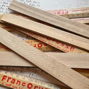 Puede incluir: Un primer plano de varias reglas de madera apiladas unas sobre otras. Las reglas son de madera clara y tienen marcas rojas. El texto "Franc Or" y "c'est un r&eacute;gal que l'on sert" es visible en las reglas.