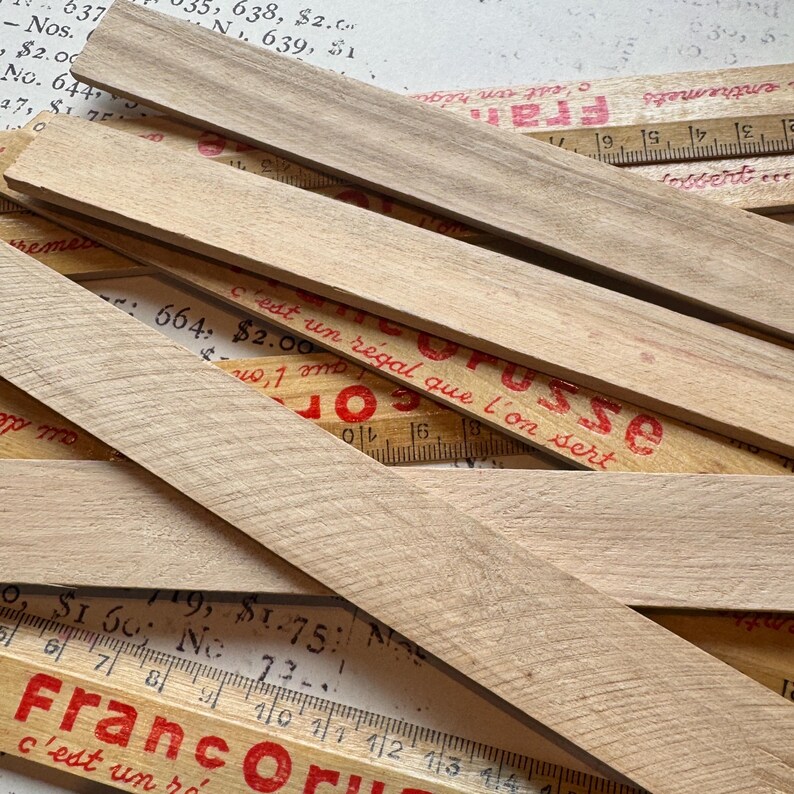 Puede incluir: Un primer plano de varias reglas de madera apiladas unas sobre otras. Las reglas son de madera clara y tienen marcas rojas. El texto "Franc Or" y "c'est un r&eacute;gal que l'on sert" es visible en las reglas.
